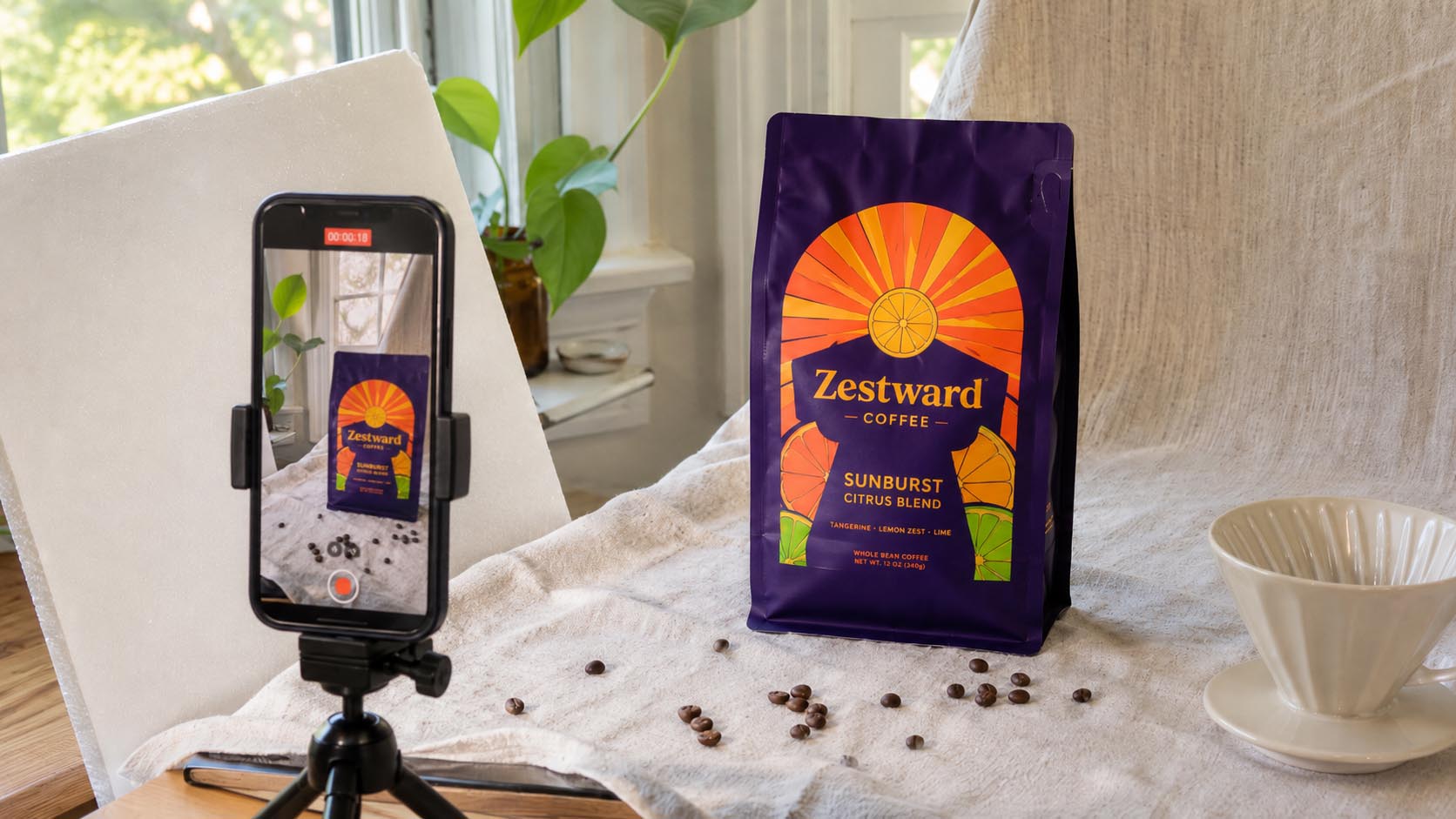 Una sesión de fotos casera de una bolsa de café en marcha: una bolsa personalizada sobre un fondo de lino junto a una ventana luminosa, con un teléfono en un trípode pequeño encuadrándola, granos y un cono de filtro como props