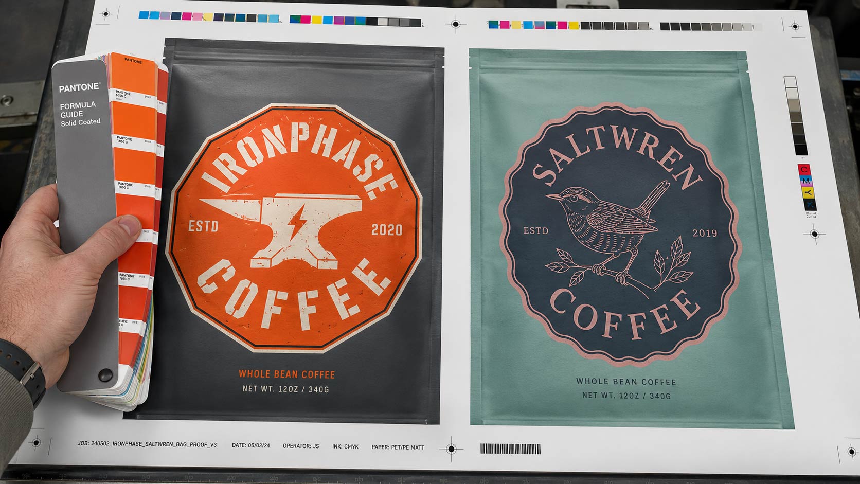 Two printed coffee bag proofs laid on a press sheet under neutral lighting while a hand checks color against a Pantone swatch book