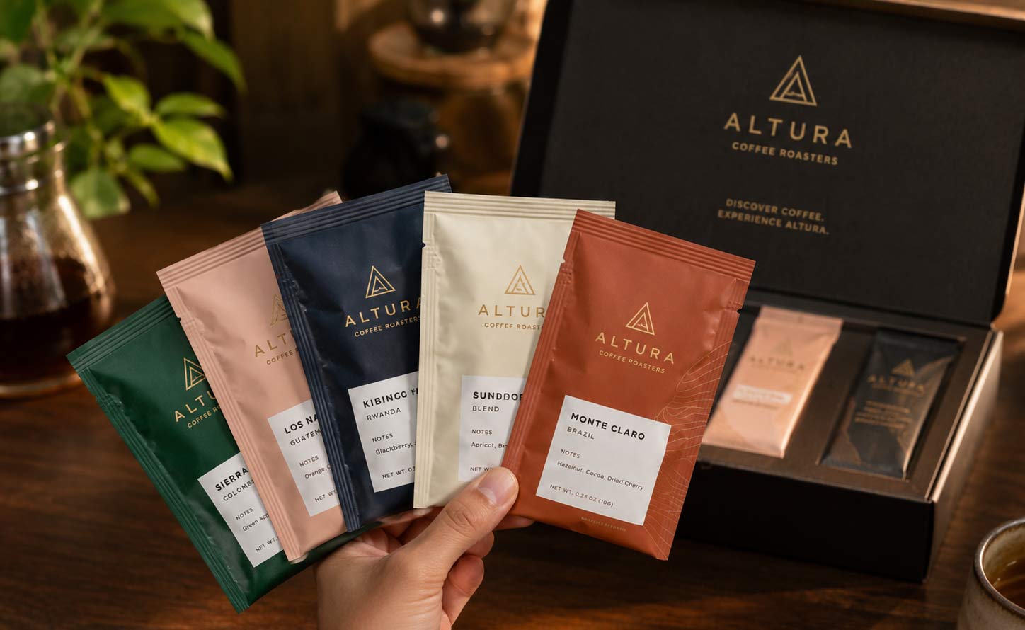 A hand holding a small fan of tri-seal coffee sample pouches with an open sample-kit box of more pouches on the table behind