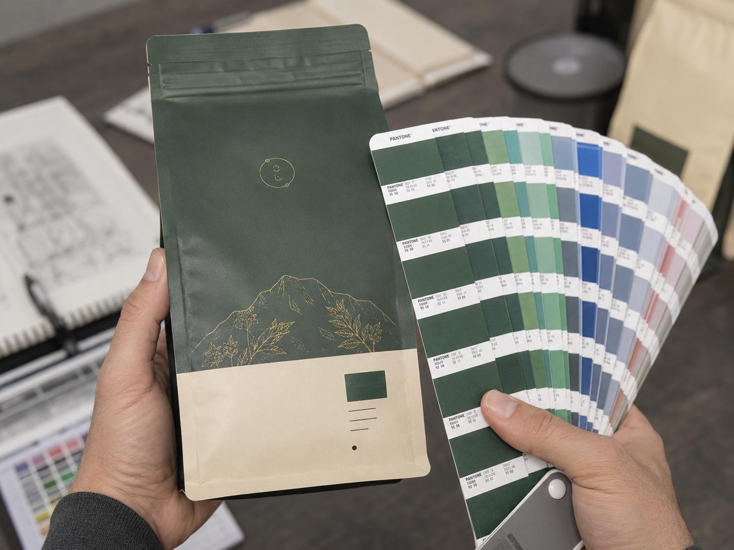 Two hands holding a printed pre-production coffee bag sample next to a Pantone fan deck under neutral light, comparing the brand color on the real bag material